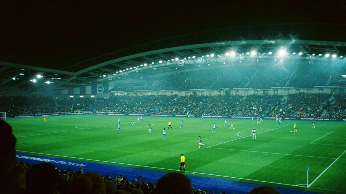 A floodlit football stadium at night during a match, with bright floodlights shining over the pitch and stands